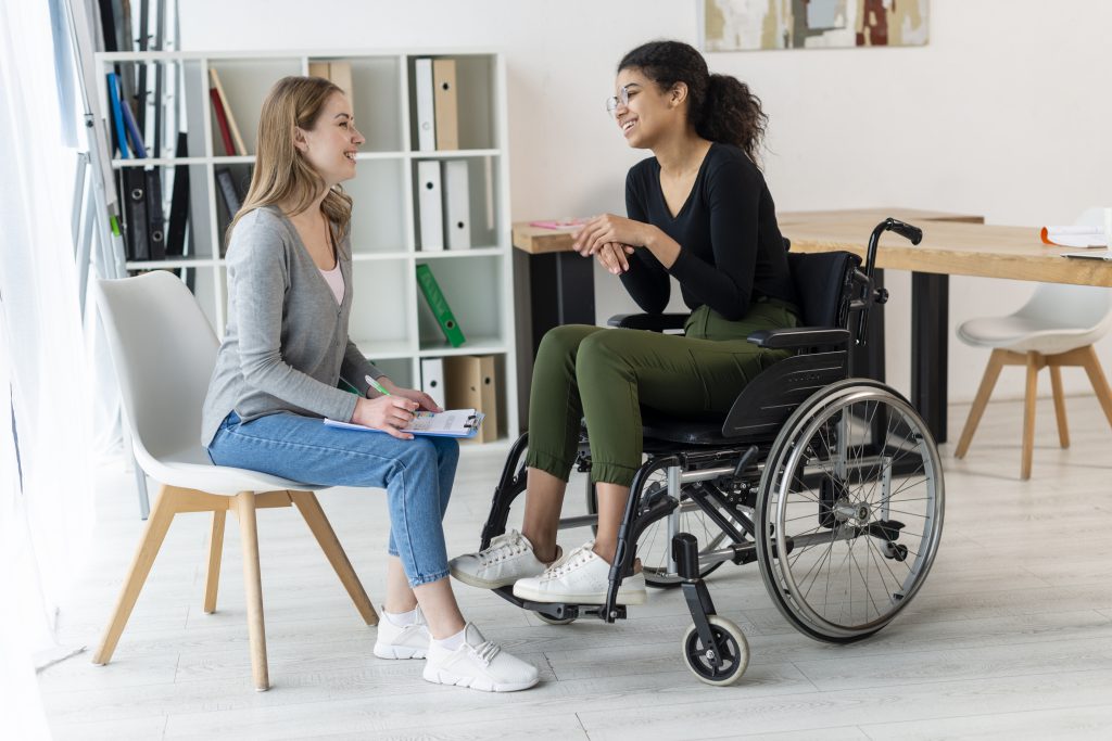 A woman holding a notepad speaking to another woman in a wheelchair